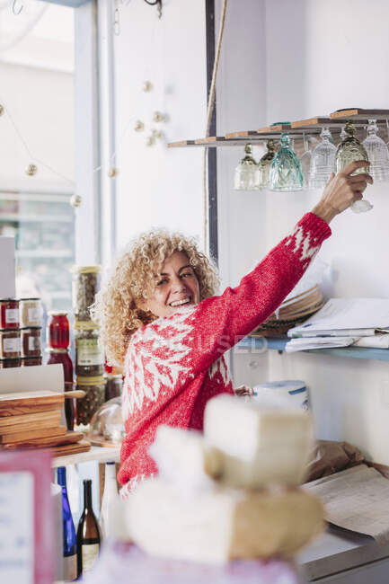 Дружелюбная взрослая женщина с кудрявыми волосами смотрит в сторону, работая в уютном местном магазине деликатесов — стоковое фото