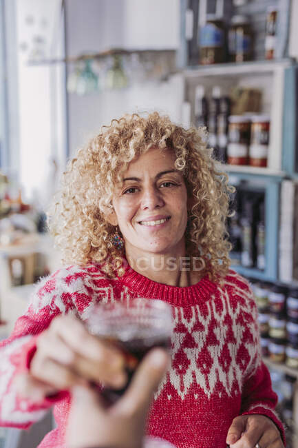 Joyeux cheveux bouclés femme blonde vendeur barman avec tasse en verre avec du vin tout en travaillant dans le magasin d'aliments délicatesse locale — Photo de stock