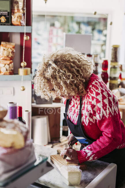 Доросла жінка в фартусі нарізає свіжий сир під час роботи в затишному місцевому продуктовому магазині — стокове фото