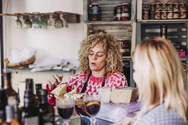 Щаслива доросла жінка з кучерявим волоссям розмовляє з клієнтом під час продажу сиру в місцевому магазині делікатесів — стокове фото