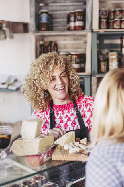 Mujer adulta feliz con el pelo rizado hablando con el cliente mientras que la venta de queso en la tienda de delicatessen comida local - foto de stock