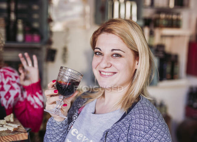 Mujer rubia feliz cliente con copa de vidrio degustación de vino mientras que las compras en la tienda de alimentos delicatessen locales - foto de stock