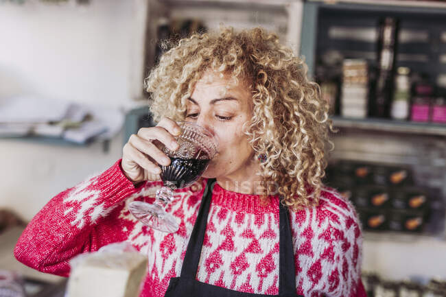 Feliz pelo rizado mujer rubia vendedora barman beber una taza de vidrio con vino mientras trabaja en la tienda de alimentos delicatessen locales - foto de stock