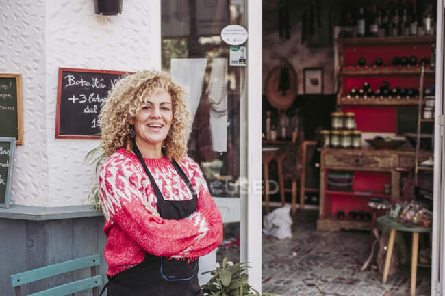 Mulher adulta positiva com braços cruzados sorrindo e olhando para a câmera enquanto estava perto da entrada da loja de alimentos local — Fotografia de Stock