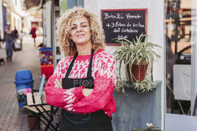 Mujer adulta positiva con los brazos cruzados sonriendo y mirando a la cámara mientras está de pie cerca de la entrada de la tienda de alimentos local - foto de stock