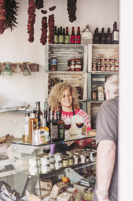 Homem sênior escolhendo comida do congelador ao comprar vinho e queijo de vendedor feminino em pequena loja aconchegante — Fotografia de Stock