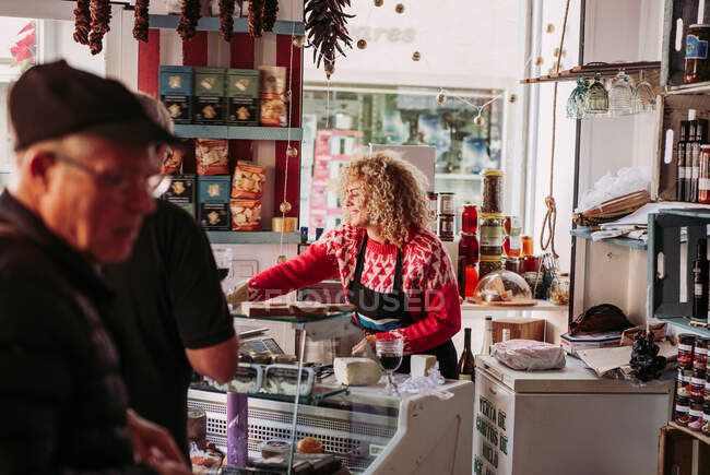 Hombre mayor eligiendo la comida del congelador al comprar vino y queso de la vendedora en la pequeña tienda de delicatessen acogedor - foto de stock