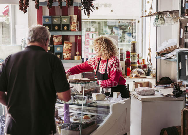 Uomo anziano che sceglie il cibo dal congelatore mentre acquista vino e formaggio da venditore femminile in accogliente piccolo negozio di gastronomia — Foto stock