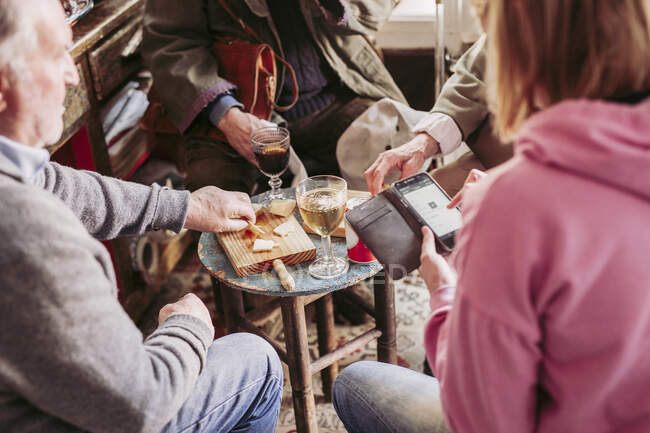 From above crop woman using smartphone while sharing company with elderly men tasting cheese and wine in cozy local food shop — Stock Photo