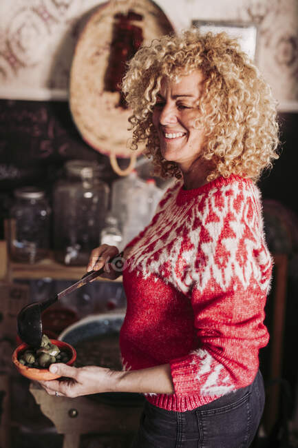 Side view of happy blonde curly woman putting ladle of olives into bowl while working in local food store — Stock Photo