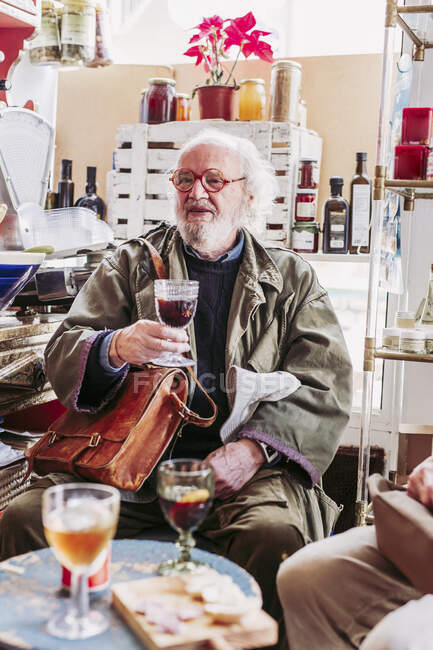 Бородатый пожилой мужчина в очках наслаждается красным вином, проводя время с друзьями в уютном местном продуктовом магазине — стоковое фото