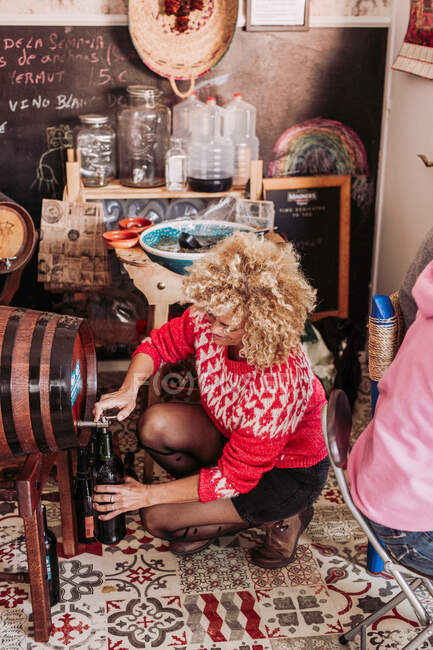 De arriba vendedor anónimo llenando taza de vidrio con vino de barril mientras trabajaba en la tienda de alimentos local - foto de stock