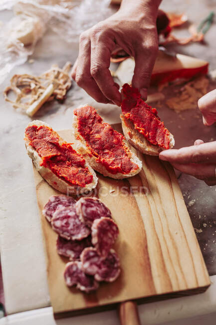 From above person holding pieces of yummy sausage and slices of bread with delicious sauce placed on wooden board in local food shop — Stock Photo