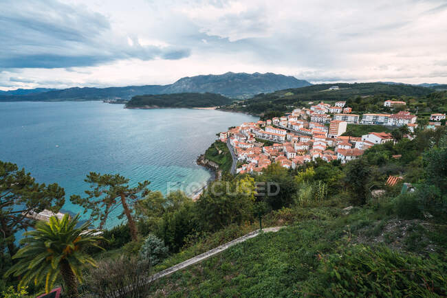 Drone view of peaceful seashore with green trees and small town with red roofs locating near blue sea and mountain ridge against blue cloudy sky — Stock Photo