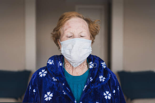 Seniorin mit roten Haaren in blauem Gewand und medizinischer Maske blickt im Krankenhauszimmer in die Kamera — Stockfoto