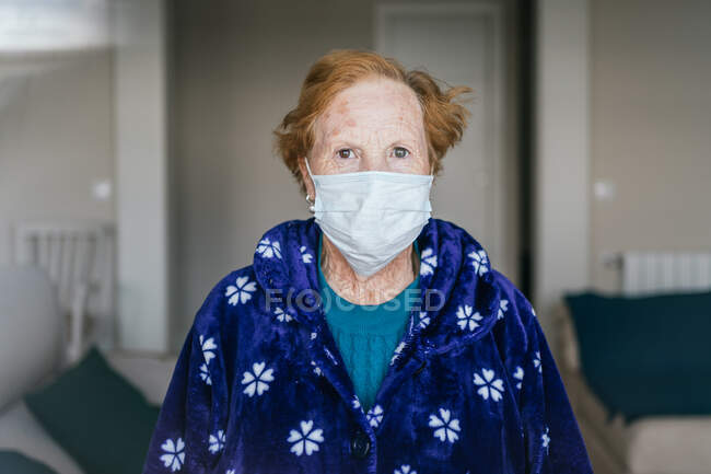 Senior female with red hair in blue robe and medical mask looking at camera while standing in hospital room — Stock Photo