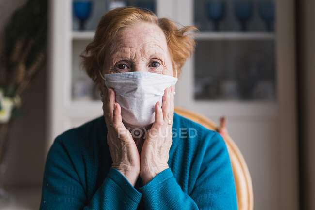 Старша жінка з рудим волоссям у блакитному халаті та медичною маскою дивиться на камеру, стоячи в лікарняній кімнаті — стокове фото