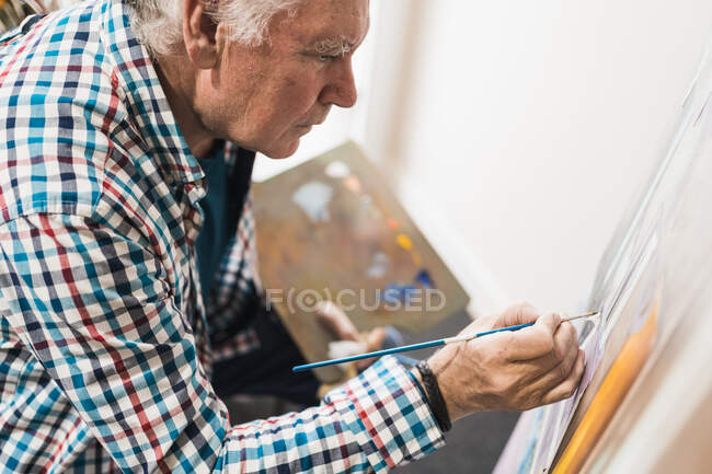 Side view of crop senior male artist in casual clothes drawing picture with paintbrush on paper on easel holding palette with paints in creative art workshop — Stock Photo
