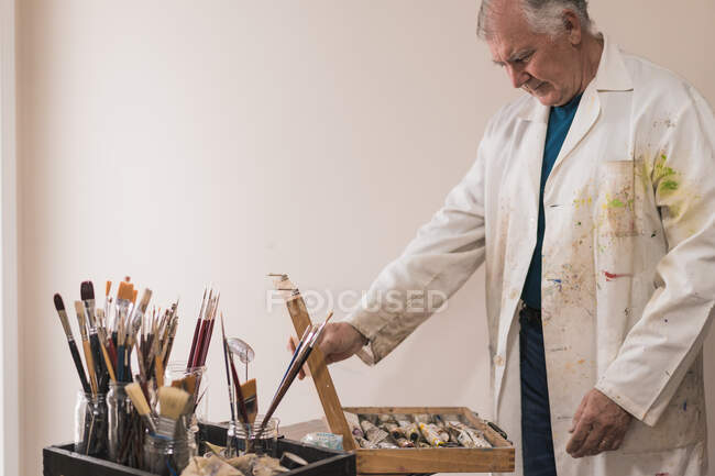 Artista di sesso maschile invecchiato in cappotto di lavoro colorato in piedi vicino al tavolo con set di pennelli e scatola di apertura con vernici durante la scelta di vernici e lavorare in studio moderno — Foto stock