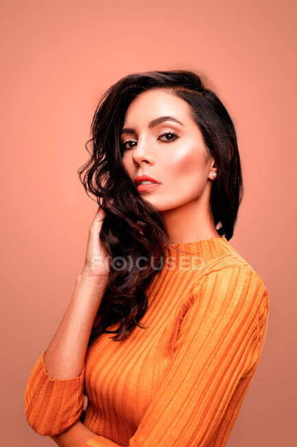 Charming confident young brunette female with long wavy hair and perfect makeup wearing casual orange sweater looking at camera while standing against pink background — Stock Photo