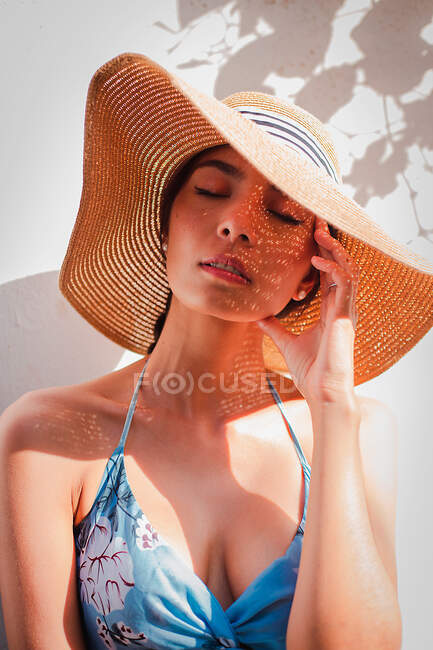 Tranquil joven hembra en vestido de sol y sombrero de paja descansando cerca de la pared blanca y disfrutando de un soleado día de verano durante las vacaciones - foto de stock