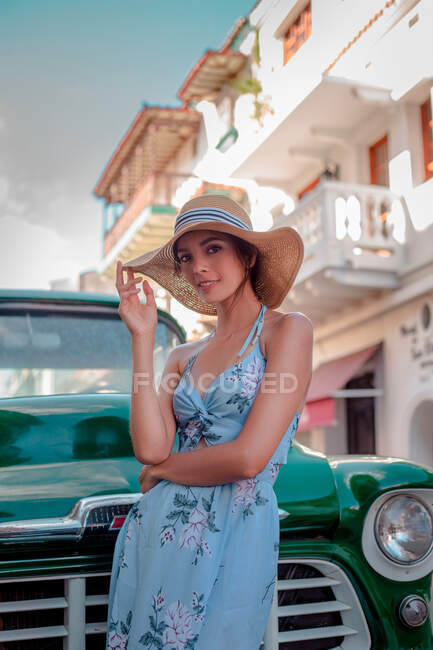 Charmante jeune femme en robe de soleil élégante et chapeau de paille regardant la caméra tout en se tenant près de voiture rétro sur la rue de la ville avec vieux bâtiment dans la journée ensoleillée d'été — Photo de stock