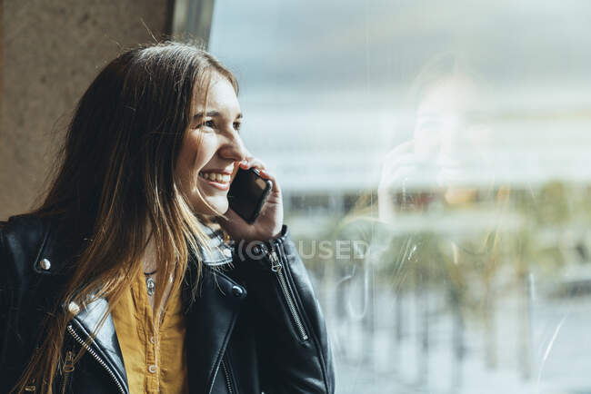 Teenager in casual clothes with suitcase and bag standing by window leaning on wall and talking on smartphone at airport — Stock Photo