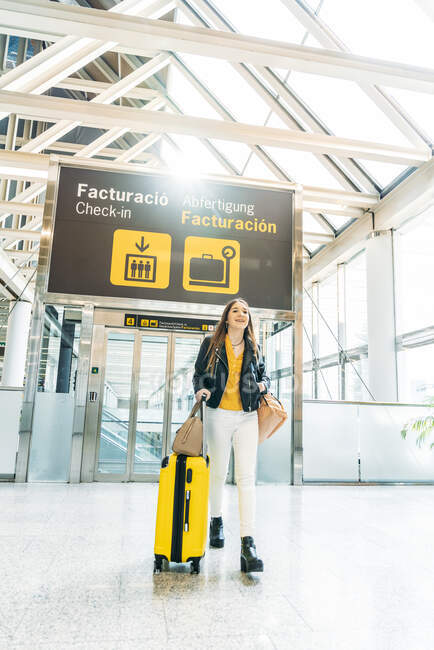 Stylish smiling teenager in yellow cap black leather jacket and yellow blouse standing with suitcase in modern airport terminal and looking at camera — Stock Photo
