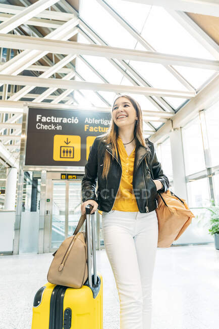 Elegante adolescente sorridente con cappuccio giallo giacca di pelle nera e camicetta gialla in piedi con valigia nel moderno terminal dell'aeroporto e guardando la fotocamera — Foto stock