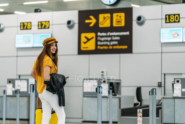 Full length of joyful teenager in fashionable outfit with luggage and passport in back pocket looking at camera and waving goodbye while standing next to check in counter at airport — Stock Photo