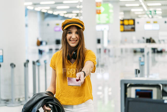 Cheerful smiling teenager in colorful stylish clothes and headphones giving passport and looking at camera while standing at check in counter in airport terminal — Stock Photo