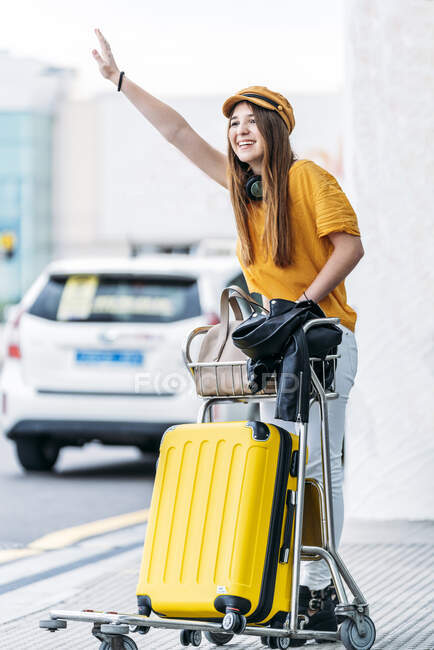Ein fröhlicher Teenager in gelber Mütze mit Kopfhörern und vollem Gepäckwagen, der an warmen Sommertagen in der Nähe der Straße vor dem großen Flughafenterminal steht und ein Taxi auffängt. — Stockfoto