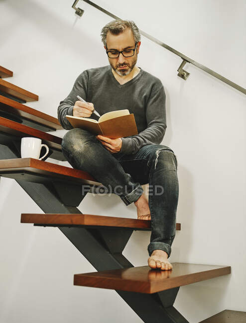 Низький кут дорослого чоловіка в окулярах відпочиває з чашкою кави на дерев'яних кроках в сучасній квартирі і пише плани в записнику — стокове фото