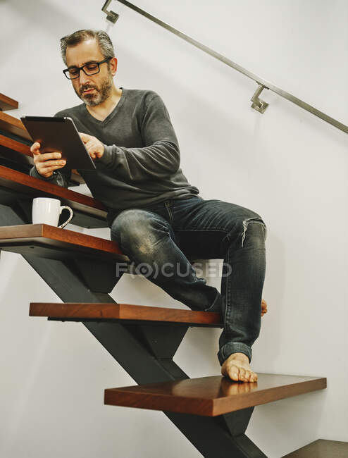 Hombre feliz en ropa casual sentado en escaleras de madera usando tableta durante el descanso en el hogar moderno - foto de stock