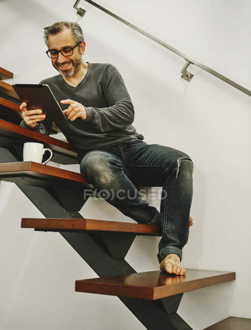Homme heureux en vêtements décontractés assis sur des escaliers en bois en utilisant la tablette pendant le repos dans la maison moderne — Photo de stock