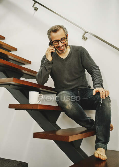 Homme riant dans des vêtements décontractés assis sur des escaliers en bois et bavardant sur un téléphone portable pendant le repos dans un appartement moderne — Photo de stock