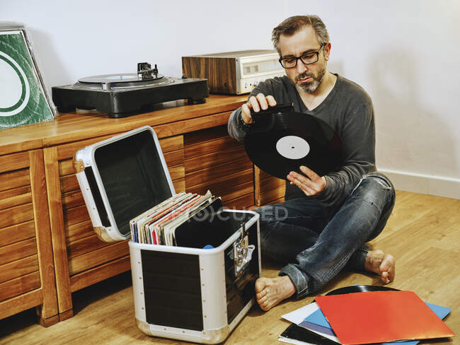 Stilvoller Mann in lässiger Kleidung und Brille sitzt auf Holzboden und pflückt Schallplatte, während er das Wochenende zu Hause genießt — Stockfoto