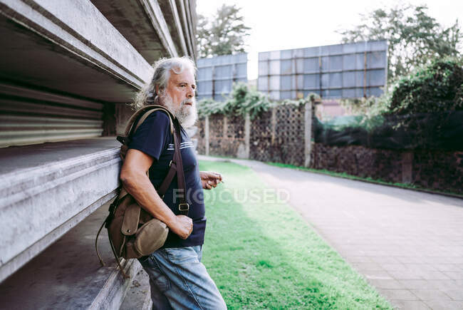 Älterer männlicher Tourist in Freizeitkleidung steht mit Rucksack in der Nähe eines Steingebäudes und raucht Zigarette auf der Straße und schaut weg — Stockfoto