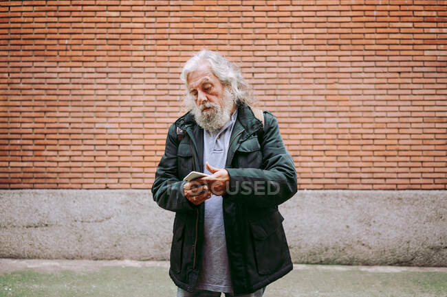 Grave uomo anziano indossa abiti casual in piedi sulla strada utilizzando smartphone sullo sfondo della costruzione di mattoni — Foto stock