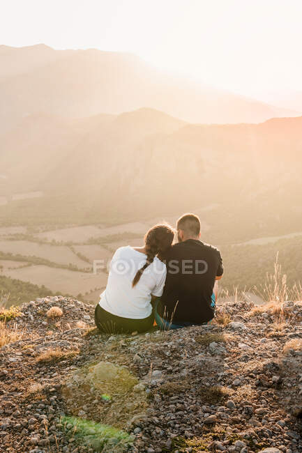 Visão traseira de casal romântico de turistas em roupas casuais sentado na borda de pedra do penhasco abraçando e desfrutando de paisagem pitoresca durante o dia ensolarado — Fotografia de Stock