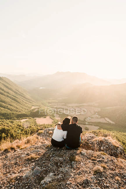 Rückansicht eines romantischen Touristenpaares in Freizeitkleidung, das auf einem steinernen Felsrand sitzt und sich umarmt und bei sonnigem Wetter die malerische Landschaft genießt — Stockfoto