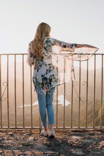 Vista trasera de la turista femenina en ropa casual de pie cerca de la valla de metal y admirando la vista del atardecer durante el paseo en el día de verano - foto de stock