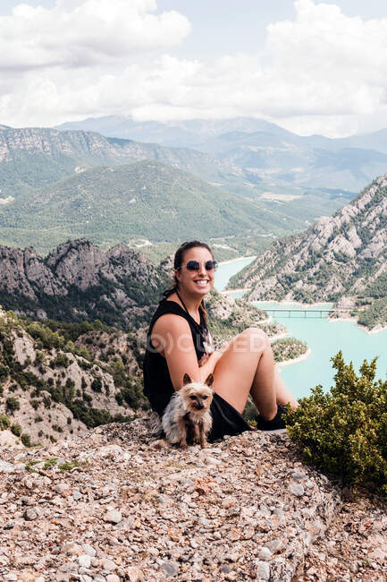 Жінка-туристка в казуальному чорному одязі і сонцезахисних окулярах сидить з собакою на скелястому пагорбі і захоплюється мальовничими пейзажами влітку — стокове фото