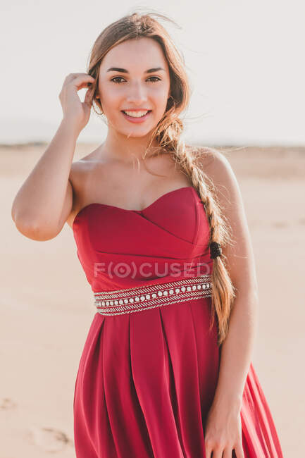 Charmante junge Frau mit langem Zopf in stylischem roten Kleid steht auf Sand und blickt in die Kamera — Stockfoto