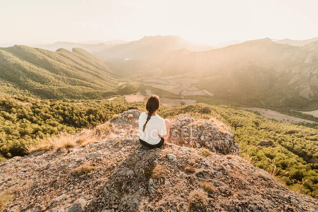 Rückansicht einer anonymen Touristin in Freizeitkleidung, die an einem sonnigen Sommertag die malerische Landschaft genießt, während sie auf steinigem Boden sitzt — Stockfoto