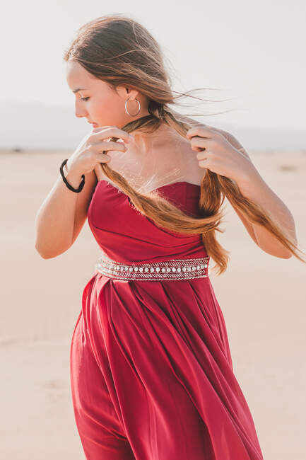 Чарівна молода жінка з довгим косичкою в стильній червоній сукні, що стоїть на піску і дивиться на камеру — стокове фото