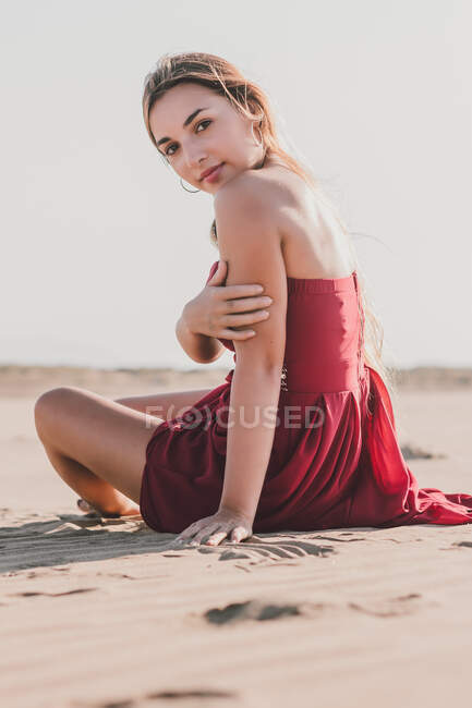 Attraktive junge Dame mit langen blonden Haaren in stylischem roten Kleid sitzt an der Küste und schaut in die Kamera — Stockfoto