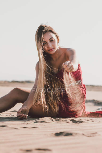 Attractive young lady with long blond hair wearing stylish red dress sitting on coast and pouring sand from hand — Stock Photo