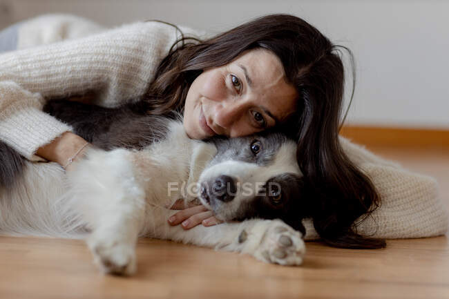 Caring female in woolen sweater hugging funny Border Collie dog while lying on wooden floor together looking at camera — Stock Photo