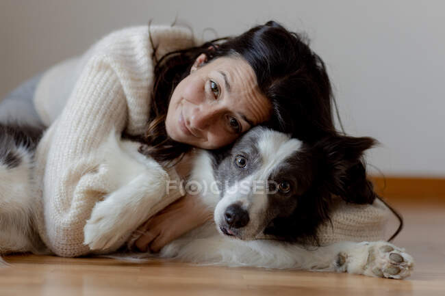 Caring female in woolen sweater hugging funny Border Collie dog while lying on wooden floor together looking at camera — Stock Photo